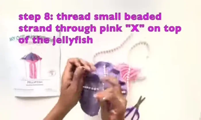 Cut the small beaded strand to fit the instruction book top to bottom. Thread it through the pink X on top of the jellyfish and connect the ends with a clasp.
